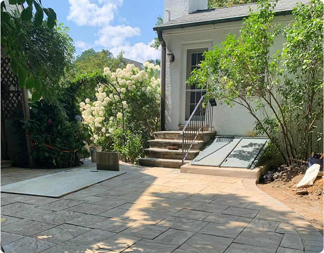 A stone patio, expertly crafted by a masonry company, leads to the back entrance of a white house with stone stairs and a slanted cellar door.