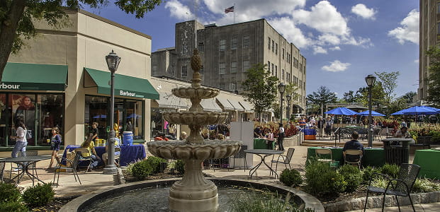 A tiered stone fountain, maintained by local masonry services, stands in a town square plaza surrounded by outdoor seating, shops, and people under blue umbrellas.