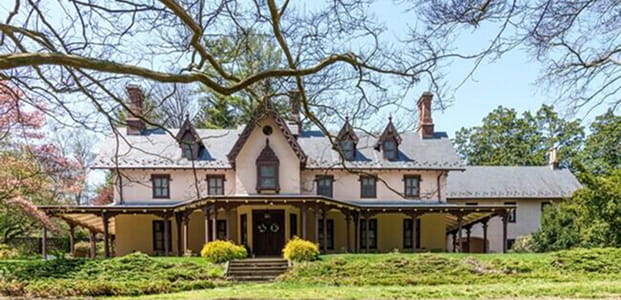 A large two-story house with a prominent wrap-around porch and steep gabled roof, framed by tree branches, representative of quality masonry service near me.