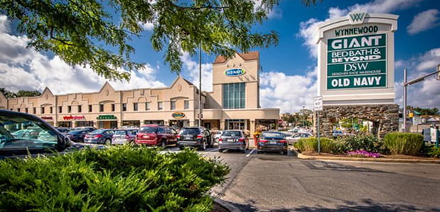 A shopping center with a large sign listing Wynnewood, Giant, Bed Bath & Beyond, DSW, and Old Navy, featuring masonry service near me, with cars parked in front of the storefronts.