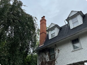White house with a red brick chimney, dormer windows, and a large tree